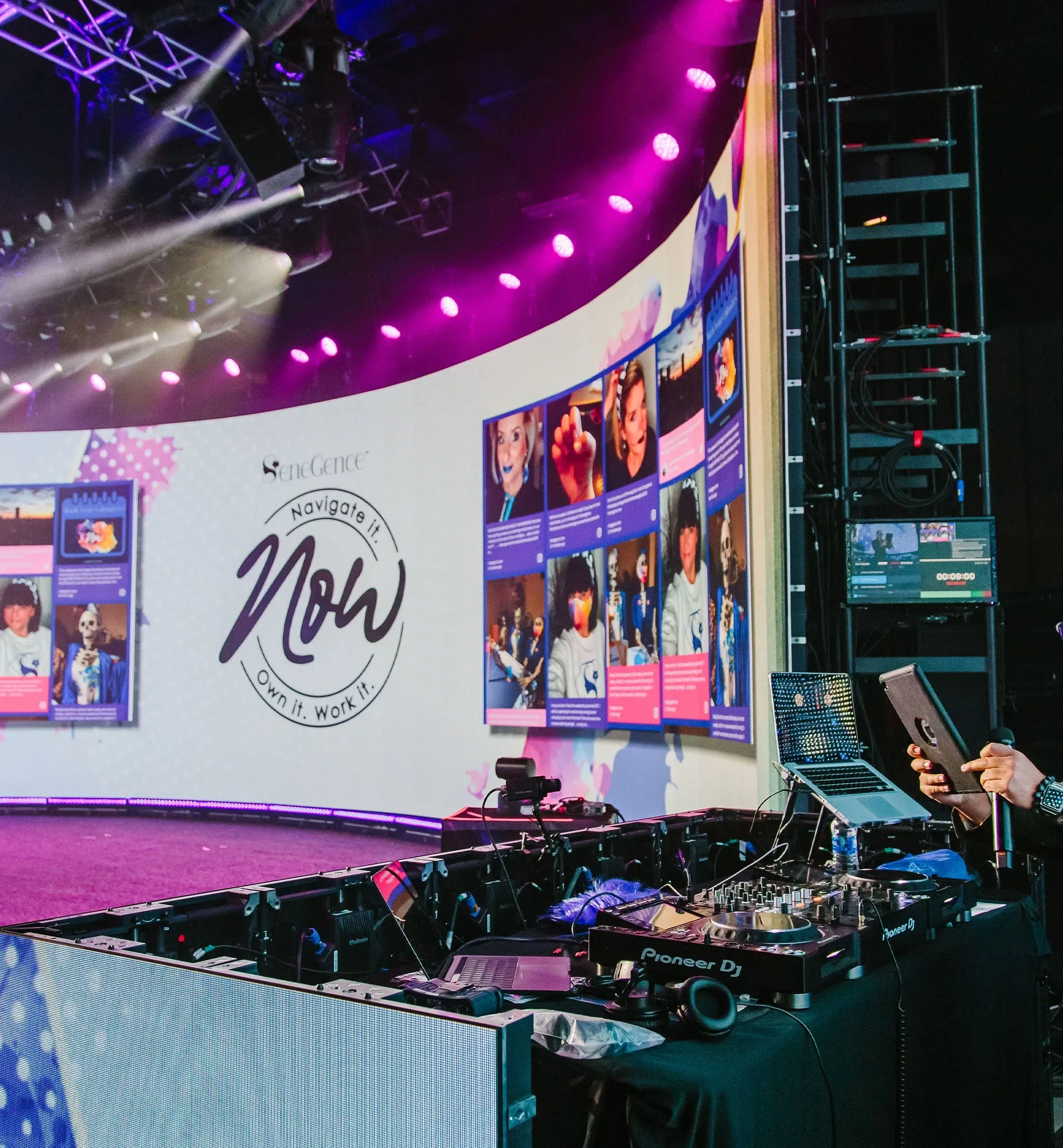 Curved LED wall showing a SeneGence Now logo and social posts behind a DJ booth with Pioneer decks