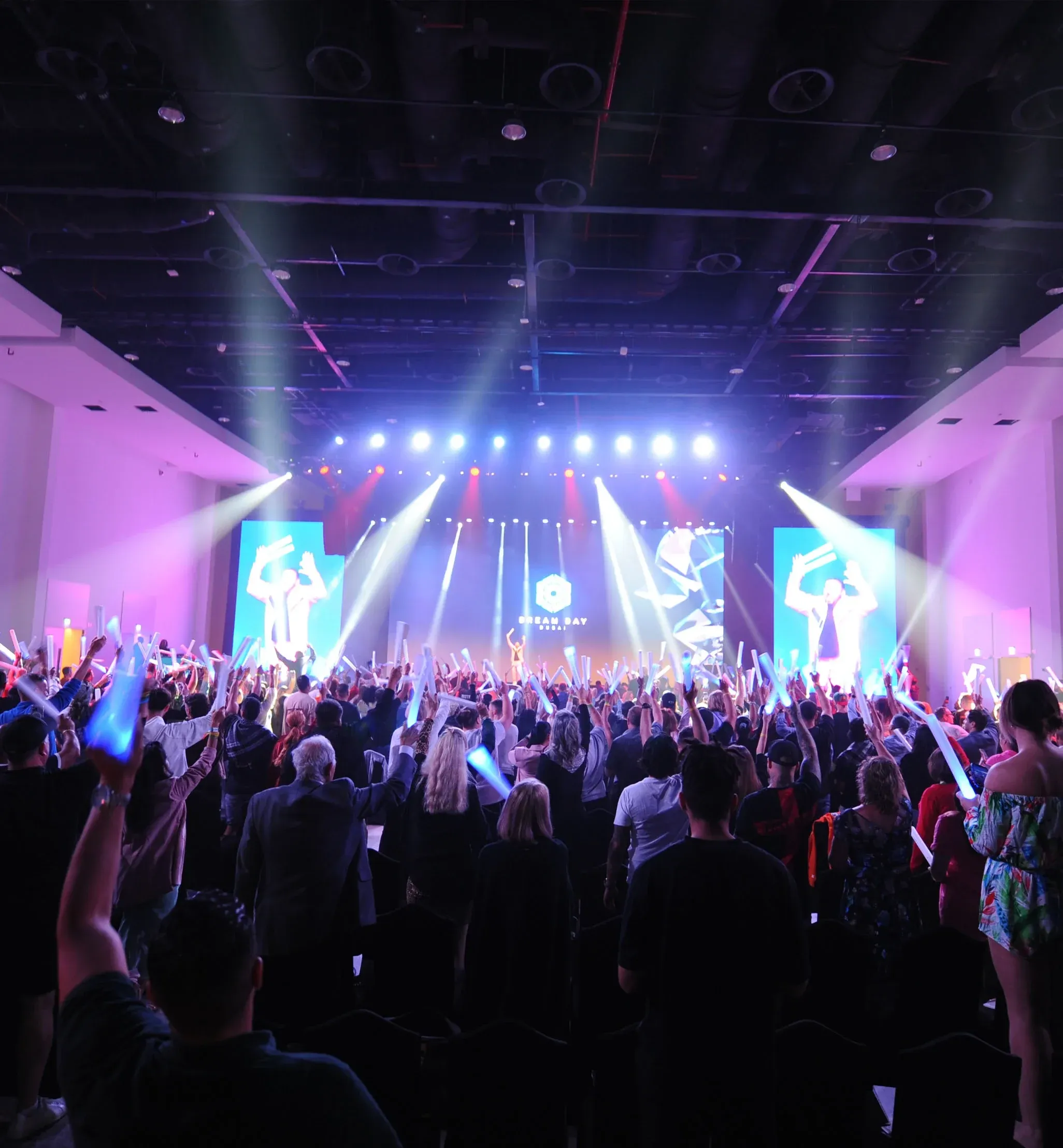Crowd waves glowing foam sticks toward a Dream Day Dubai stage with beam lights and video screens