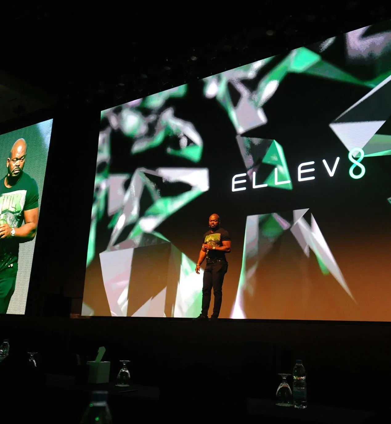 Speaker stands before a large ELLEV8 video screen with green geometric graphics and side IMAG