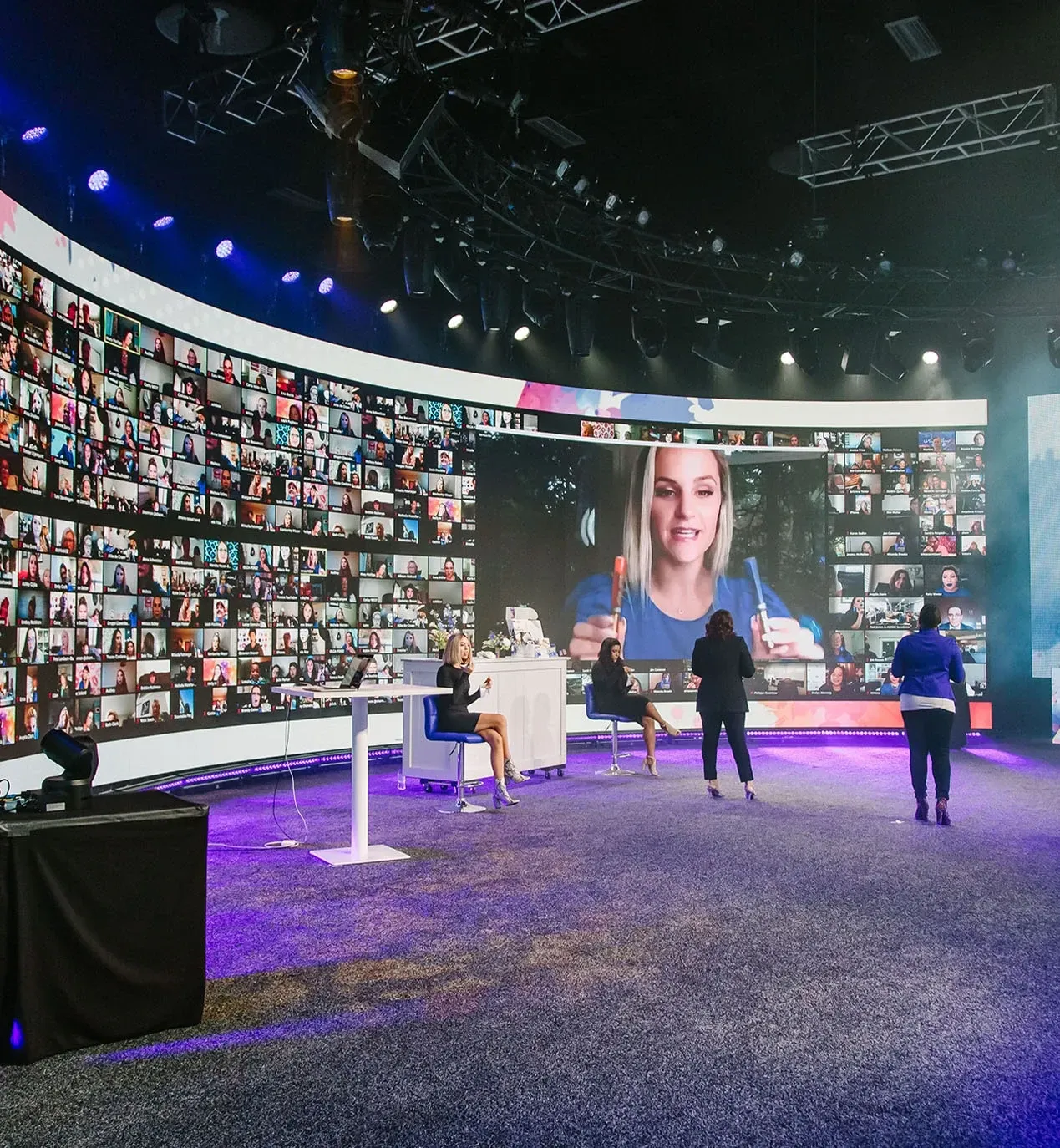 Hosts on a broadcast set before a curved wall of virtual attendees and a remote presenter holding products