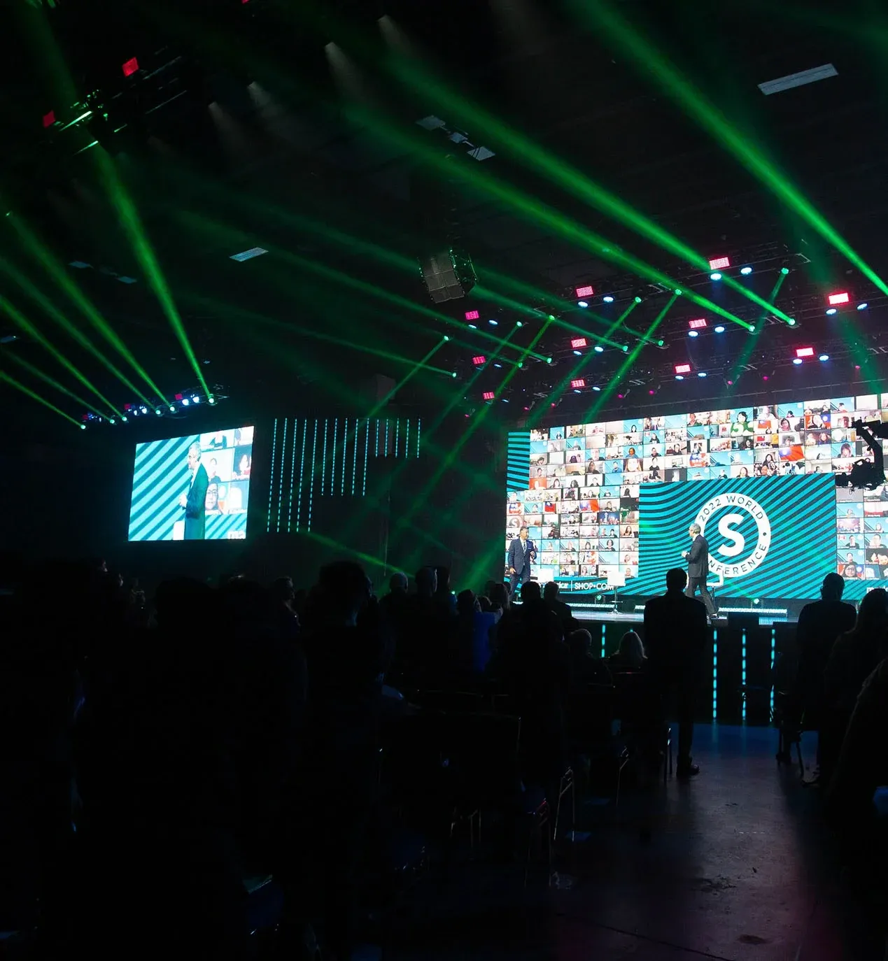 Green beam lights sweep over a dark audience facing a 2022 World Conference stage and IMAG screen