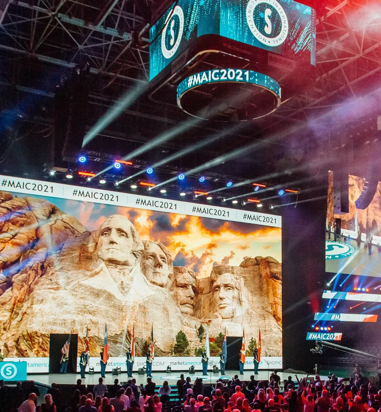 Flag bearers stand on stage before a Mount Rushmore image under #MAIC2021 banners and a center-hung screen