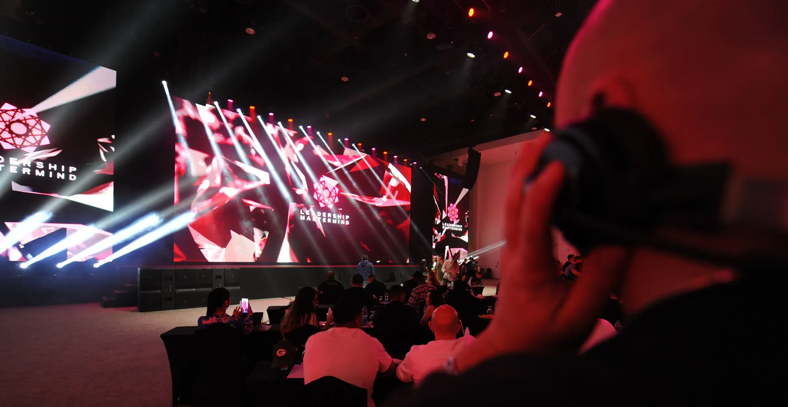 Seated attendees face red Leadership Mastermind screens crossed by white beam lights, a videographer in the foreground