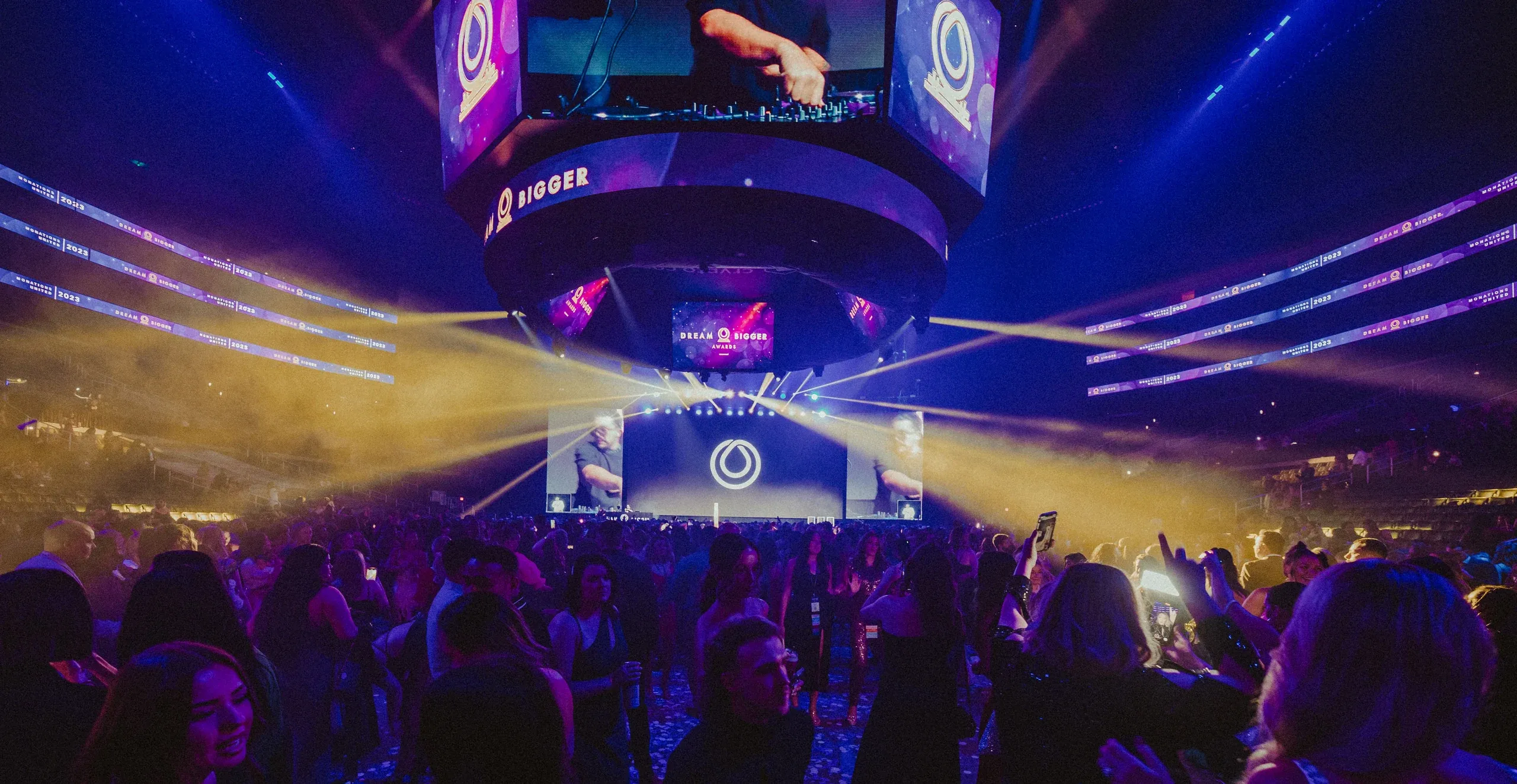 Dancing crowd on the arena floor as yellow beams sweep beneath a Dream Bigger Awards center-hung screen