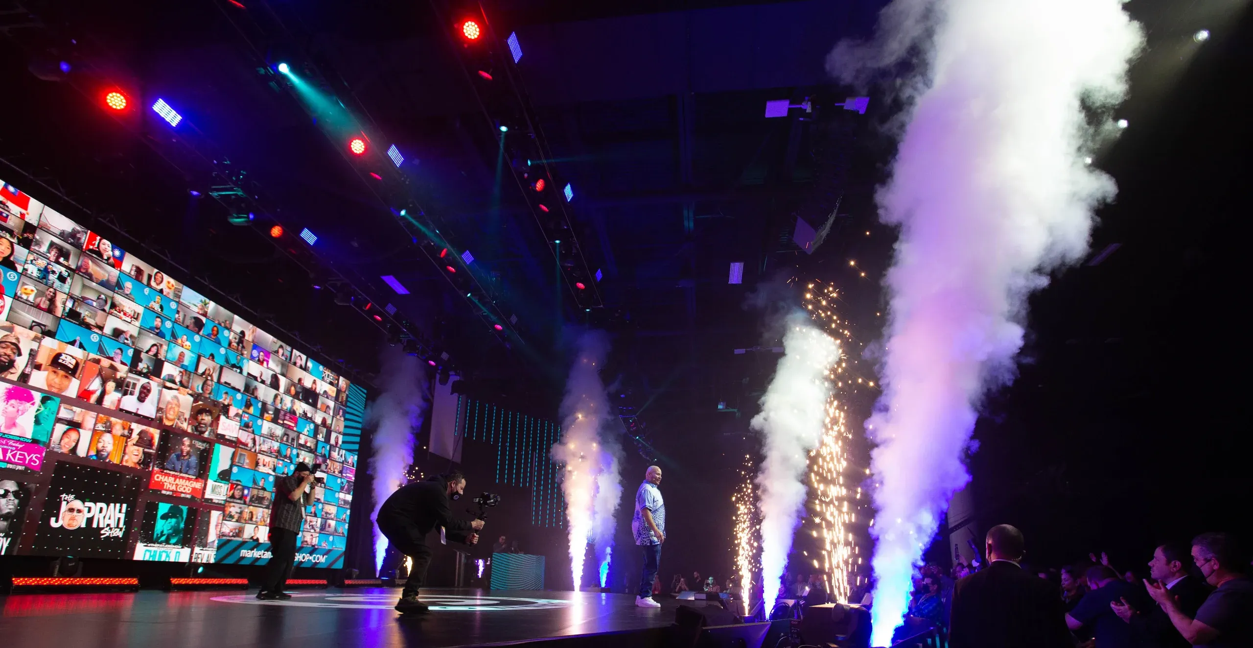 Performer walks on stage between CO2 jets and sparkler fountains as camera operators film beside a virtual-attendee wall