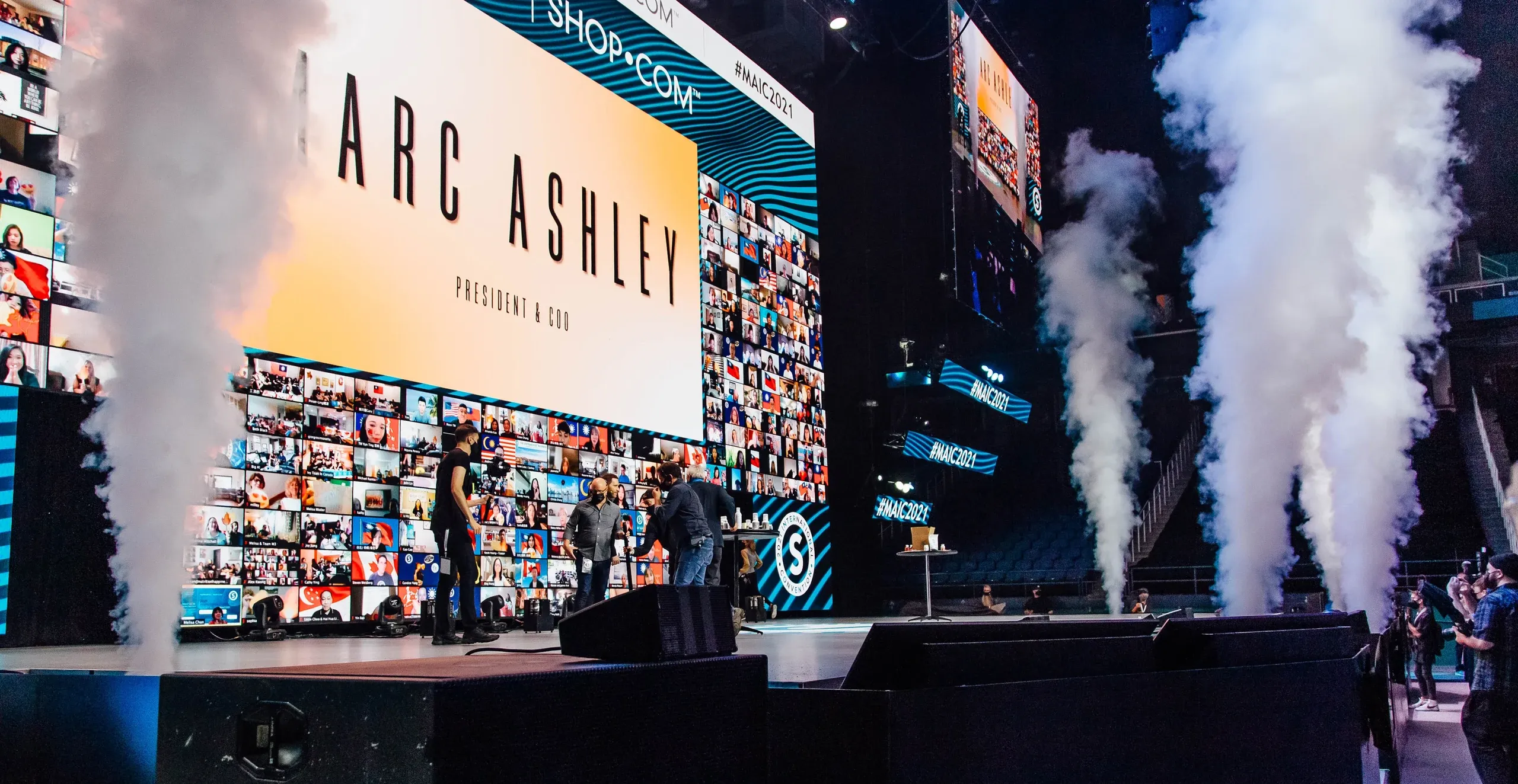 CO2 jets blast beside a stage where crew prepare in front of an executive title graphic and virtual-attendee walls