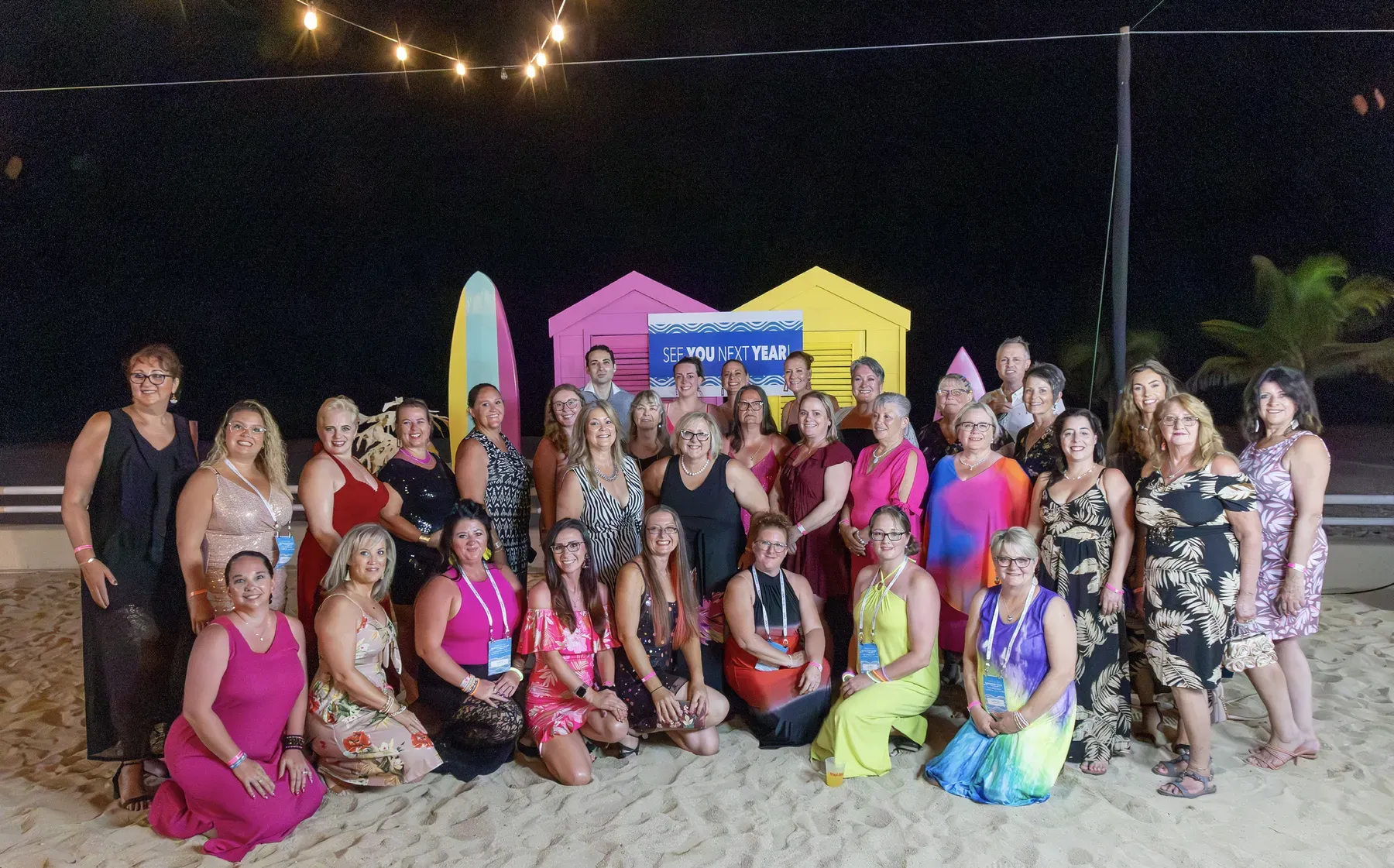 Color Street stylists in bright dresses pose on the sand in front of colorful beach huts