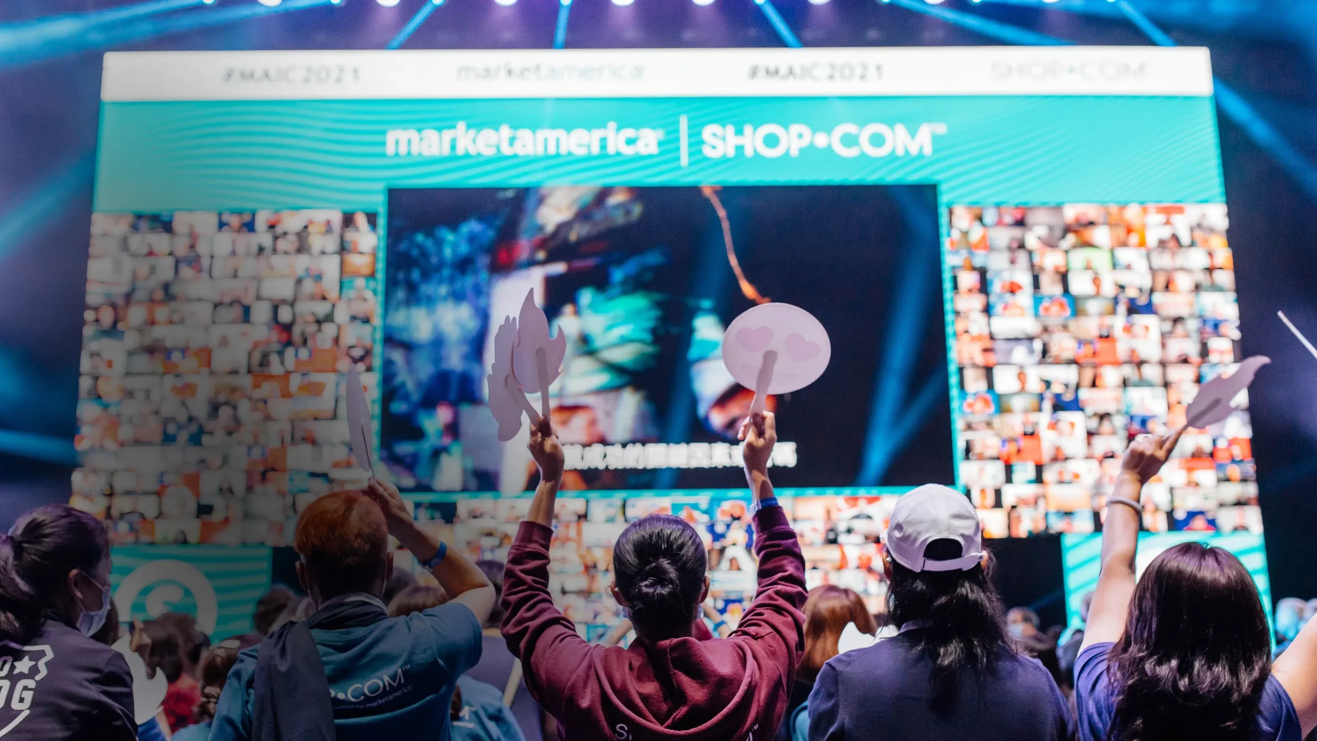 Attendees raise emoji paddles toward a Market America SHOP.COM screen framed by walls of virtual attendees
