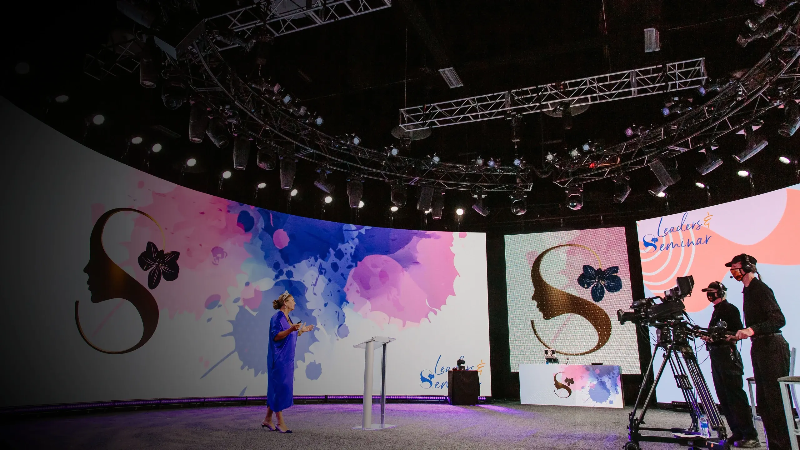 Presenter speaks on a curved LED set with SeneGence and Leaders Seminar graphics as camera operators film
