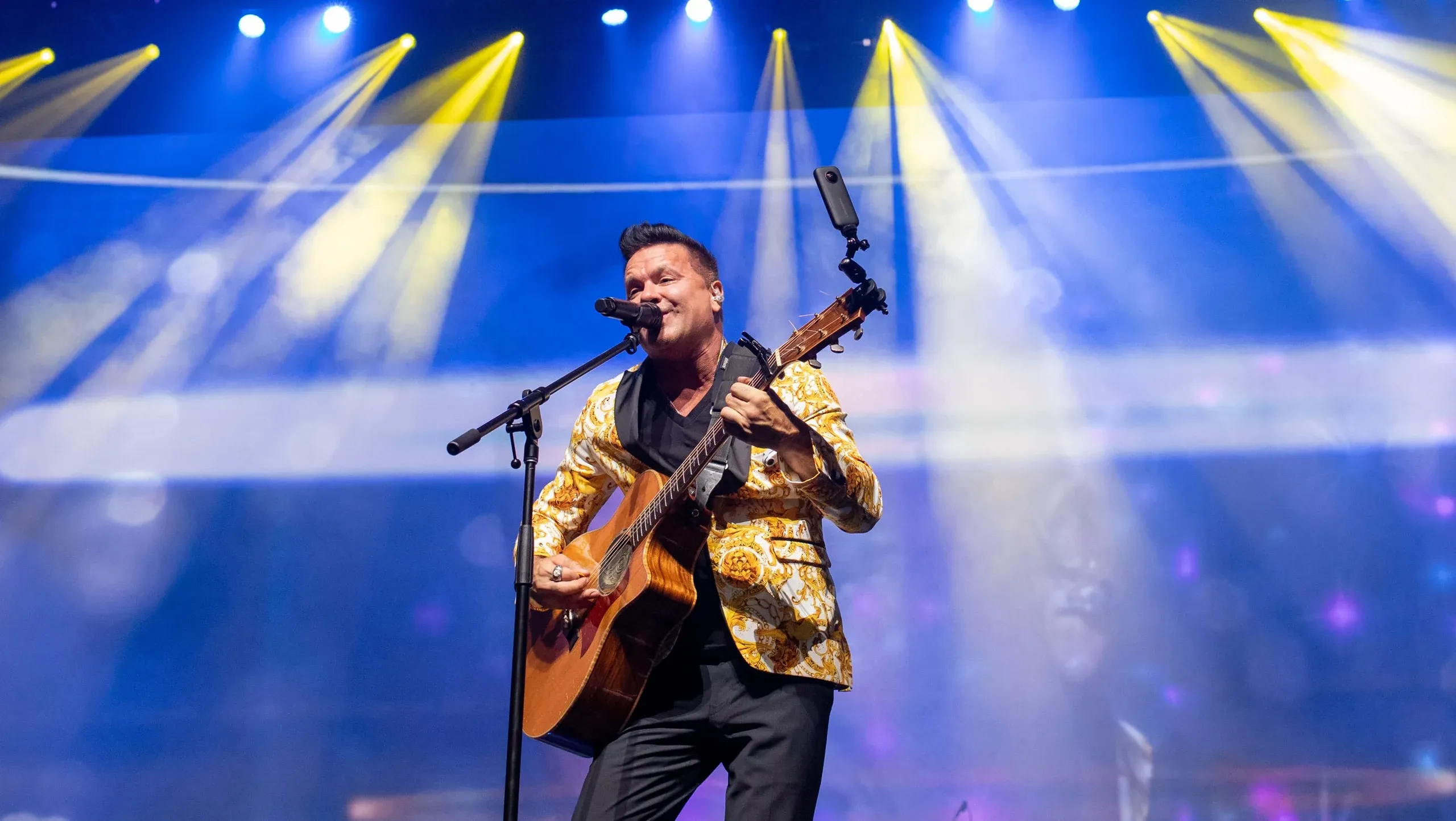 Singer in a gold patterned jacket plays acoustic guitar at a microphone under yellow and blue beam lights