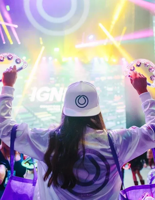 Attendee in a Monat-logo cap and jacket raises tambourines toward a brightly lit IGNITE stage
