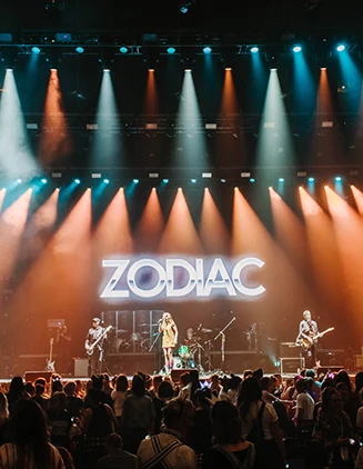 rock band plays on stage lit by orange and blue lights with the band's name, Zodiac, above them